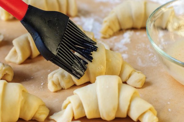 Croissants making and brushing with egg on white table with flour and baking paper