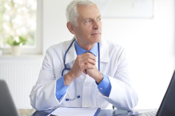 Thinking male doctor sitting in at desk behind computers in the consulting room.