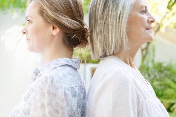 Close up family side portrait of an adult daughter with her mature mother standing back to back against each other while standing in a home garden together, otudoors.