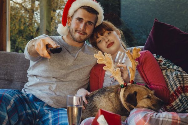 Young couple at home with their pet dog at Christmas time. They are all cuddled up on the sofa at home, watching television.