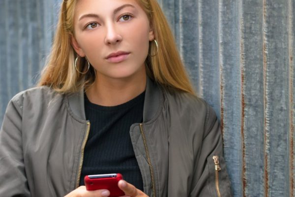 Outdoor portrait of pretty sad thoughtful blonde girl teenager female young woman using a red smart cell phone wearing green bomber jacket and aviator sunglasses