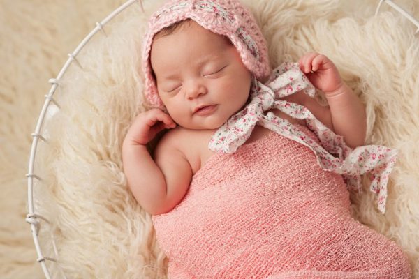 A portrait of a five week old newborn baby girl wearing a pink bonnet. She has a subtle smile and is peacefully sleeping in a wire basket.
