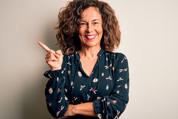 Middle age beautiful woman wearing casual dress standing over isolated white background with a big smile on face, pointing with hand and finger to the side looking at the camera.