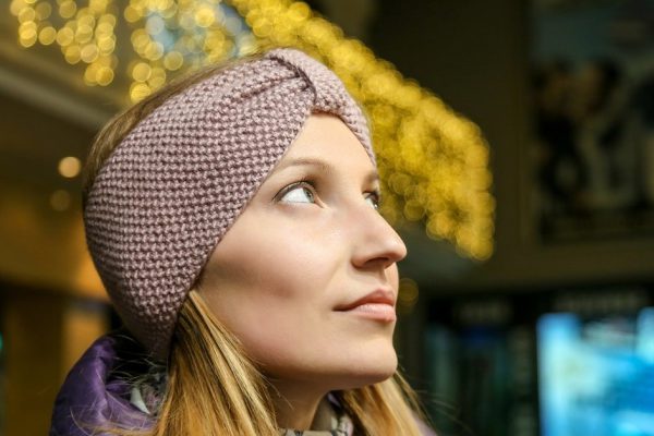 Portrait of young pretty woman in beautiful knitted woolen headband outdoors at cold autumn day. Evening bokeh lights on background. Handmade craft fashion clothes.