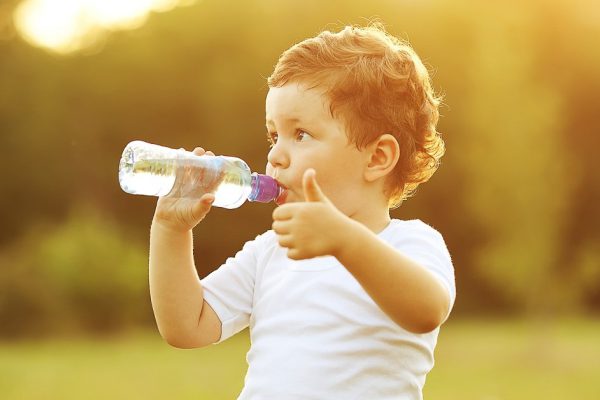 baby boy with brown hair drinking water in the park, holding plastic bottle and showing thumb up. outdoor shot