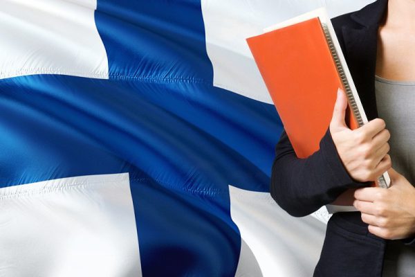 Learning Finnish language concept. Young woman standing with the Finland flag in the background. Teacher holding books, orange blank book cover.