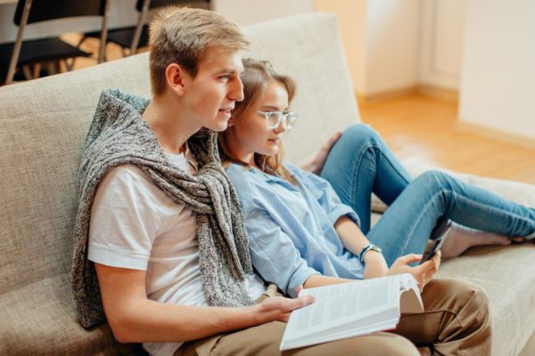 young beautiful couple sitting on sofa at home, leisure together, man reading book, woman using phone