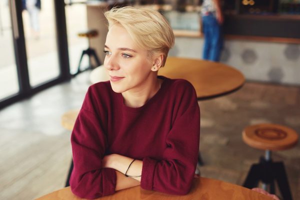 Young blonde female hipster with trendy short haircut looking into the window feeling calm during recreational time while waiting for cup of coffee in cafe sitting near copy space for your advertising