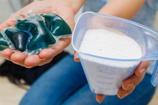 Woman in bathroom doing laundry, choosing the best detergent. Female holding powder container and gel pods for washing clothes. Household duties.