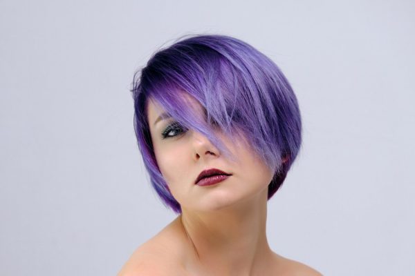 A beautiful, sexy girl with purple hair and a short haircut sits in the middle of the photo with a white background and grimaces. She's naked and visible shoulders.