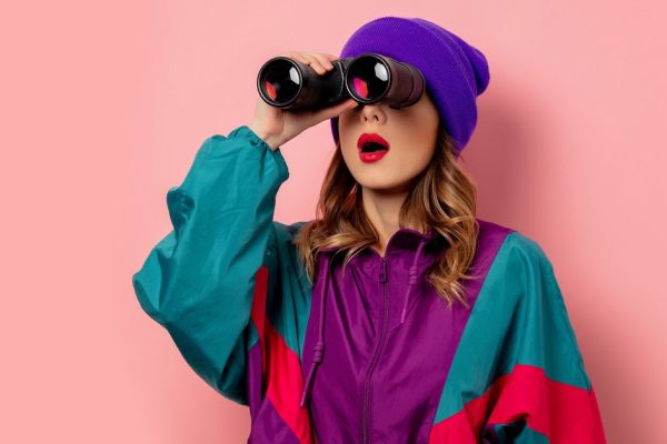 Young woman in purple hat and 90s clothes with bonoculars on pink background
