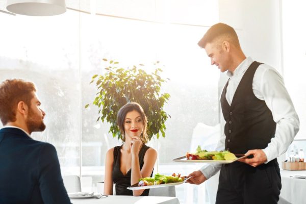 Happy Couple In Love Having Romantic Dinner In Luxury Gourmet Restaurant. Waiter Serving Meal. People Celebrating Anniversary Or Valentine's Day. Romance, Relationship Concept. Healthy Food Eating.