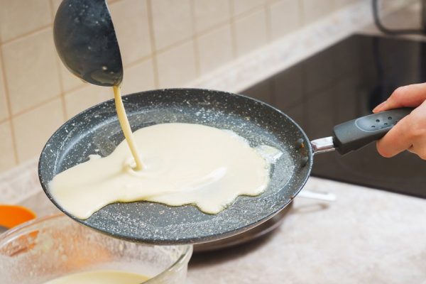 The process of making traditional thin pancakes in a frying pan. Woman pours the batter into a thin pancake pan. Soft focus and appliances in the backgrounds.