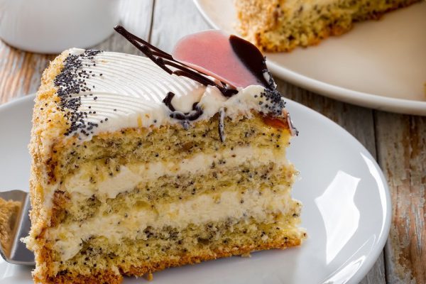 sponge cake with poppy seeds layered with cream with a cut out piece on cake shovel on white plate close-up with jug of milk, cup with black tea on saucer on peeling paint planks, view from above