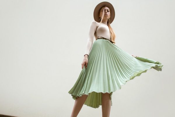 Young red hair girl in hat, dressed in beige blouse and turqoise pleats skirt, on light street backgraund.  Fashion and stylish concept.