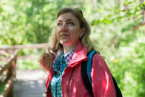 A woman in a tourist jacket and backpack admires the nature in a forest park. Summer sunny day
