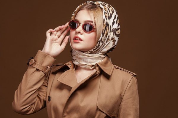 Fashion portrait of young stylish woman in trendy trench coat, leopard print headdress and fashion sunglasses over beige background.