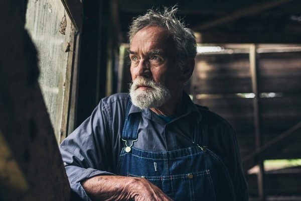 Elderly man staring out of a rustic wooden window in an old rural barn or house with a thoughtful serious expression