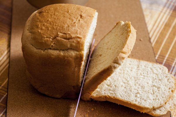 Slicing a loaf of bread. A large knife cuts thin pieces of white bread from a rectangular loaf. View from above