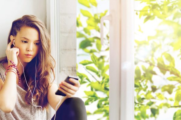 people, technology and teens concept - sad pretty teenage girl sitting on windowsill with smartphone and earphones listening to music