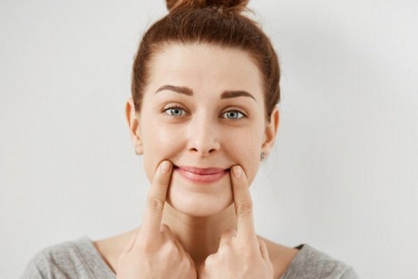 Headshot of young Caucasian woman making fake smile with her fingers stretching the corners of her mouth. Portrait of student girl trying to stay positive after failing final exams at university