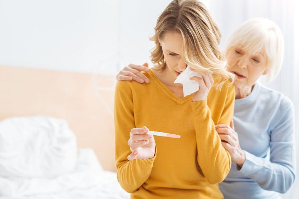 Crying woman. Young worried emotional woman sitting on the bed and crying when seeing the results of her pregnancy test while a kind supporting attentive grandmother touching her shoulders